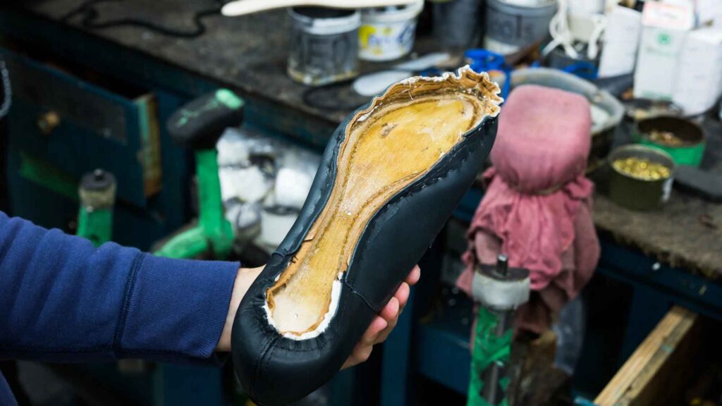 A person holds a shoe in progress with the sole exposed, surrounded by tools and materials, applying insole glue meticulously in a workshop