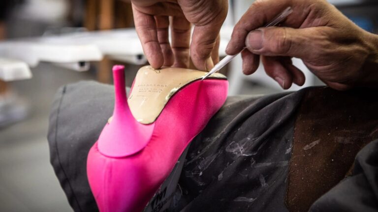 A person is holding a bright pink high-heeled shoe. The focus is on their hands and the shoe, suggesting detailed craftsmanship