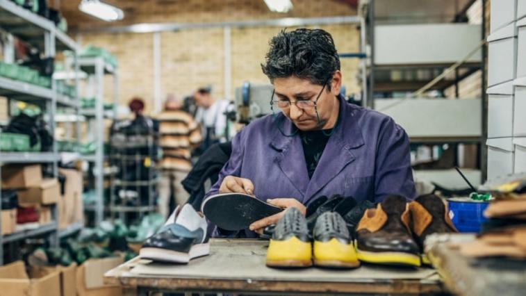 A person wearing a purple lab coat inspects a shoe sole at a workbench, focusing on bonding insoles in the workshop