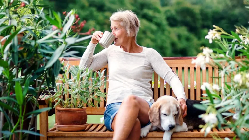 An older woman sits on a bench garden drinking from a mug, with her other hand petting a beagle sitting next to her. There are potted plants beside her, and a green landscape is in the background